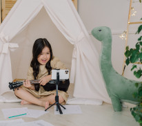 Ein Mädchen sitzt vor einem weißen Zelt in ihrem Kinderzimmer und spielt auf einer Gitarre, daneben steht ein großer grüner Dino aus Stoff.