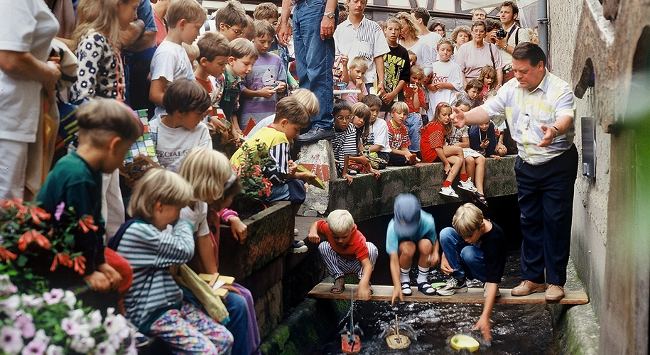 Drei Kinder lassen ihre selbstgebastelten Bötchen bei der Gerberbachregatta zu Wasser, umringt von zahlreichen weiteren Kindern