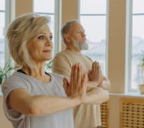 Ein älteres Paar übt Yoga in einem Raum mit vielen Fenstern.